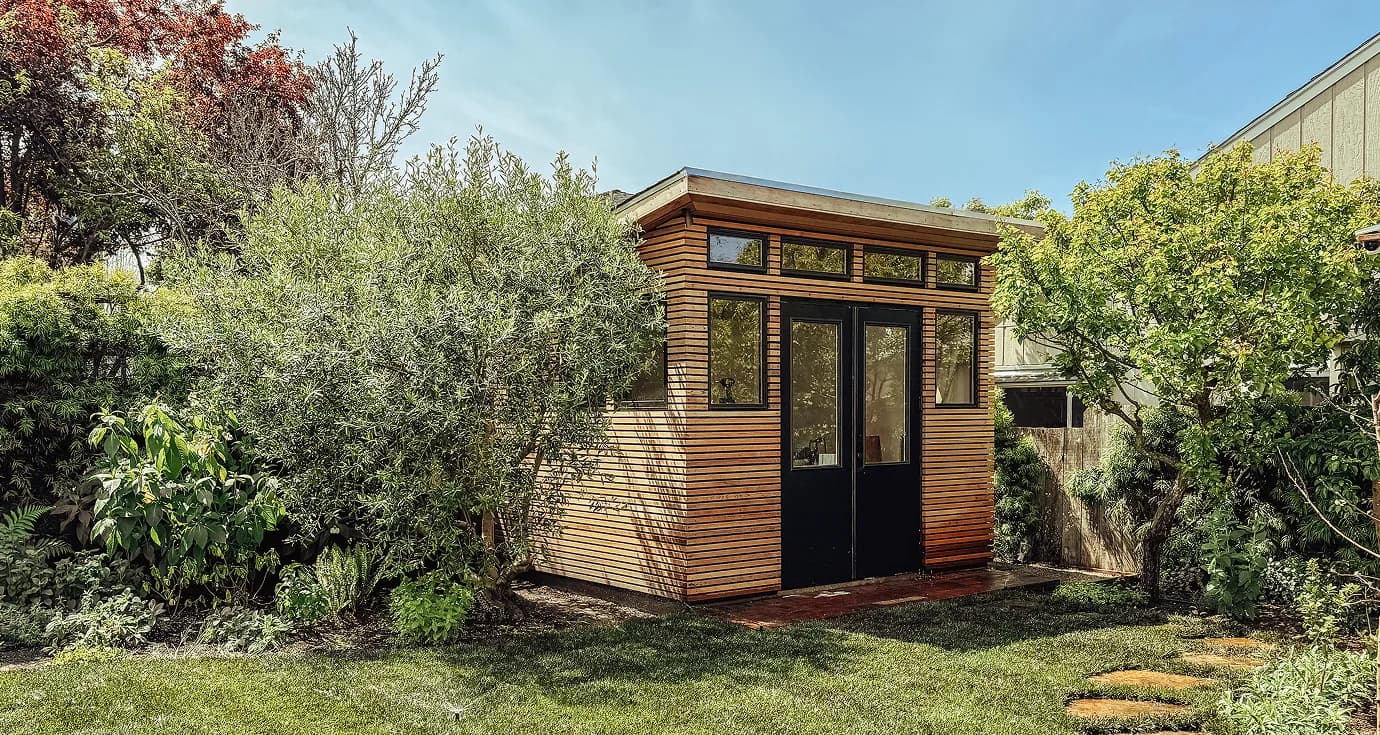 The finished building in the garden, clad in cedar with black-framed glass doors and a row of clerestory windows above